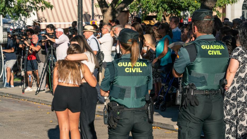 Dos agentes de la Guardia Civil observan a dos chicas abrazadas durante el minuto de silencio por el asesinato de un menor, en la puerta del Ayuntamiento de Mocejón, a 18 de agosto de 2024, en Mocejón, Toledo, Castilla-La Mancha (España). El Ayuntamiento de Mocejón ha decretado tres días de luto oficial por el asesinato en la mañana de hoy de un menor de 11 años, que se encontraba jugando al fútbol junto a otros niños de la localidad en el campo de fútbol municipal 'Ángel Tardío' cuando fue agredido con un objeto punzante.
18 AGOSTO 2024;ASESINATO;MENOR;NIÑO APUÑALADO;TOLEDO;CRIMEN;MINUTO DE SILENCIO;GUARDIA CIVIL
Juan Moreno / Europa Press
18/8/2024